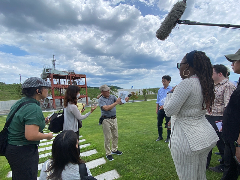 Outdoors, several participants in the Japan training program are gathered around a Japanese man explaining disaster prevention equipment.