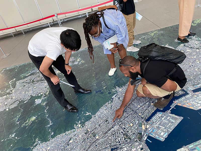 A male participant in the Japan training program is pointing at a large map printed on the floor, while two other men observe him.
