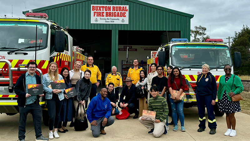 At an Australian fire station, 19 people—including trainees and firefighters—are posing for a commemorative photo in front of a fire truck.