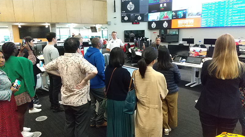 The backs of training participants touring a facility with numerous monitors.