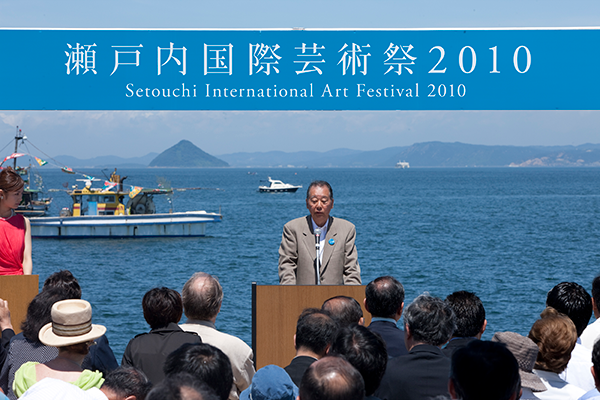 海を背景に福武氏が壇上にいる。聴衆の後ろ姿が20名ほど写っている。