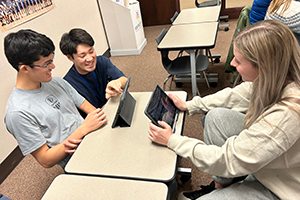 教室で3人の人物が机の周りでタブレットを見ている