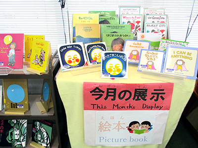 黄色いクロスで覆われて、5月の月展示のテーマの「絵本」が多数飾られた図書館の一角の机。「はじめてのおつかい」や「おつきさまこんにちは」などのタイトルが並んでいる。」
