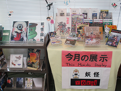 黄色いクロスが敷かれたテーブルの上に7月の展示のテーマの「妖怪」についての書籍が多数飾られた図書館の一角。