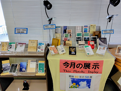 黄色いクロスが敷かれたテーブルの上に8月の展示のテーマの「戦後80年の50冊」についての書籍が多数飾られた図書館の一角。