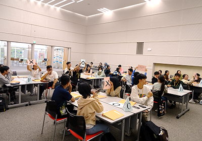 広い会議室のような室内で、AからFまで番号の書かれた三角錐型の目印が置かれた机に生徒が四人ずつ座り、〇✖の書かれたパネルを掲げて研修に参加している