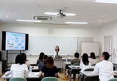 女性が教壇に立って、教室左前にモニターを置いて授業をしている。
