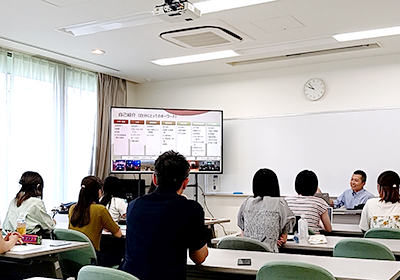 男性が教壇で、教室左前に置かれたモニターを見ながら座って授業をしている。