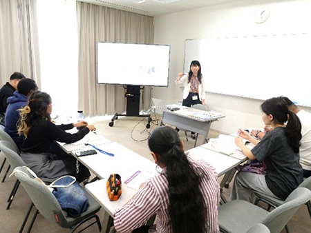 教室で1人の女性がホワイトボードの前に立っていて、5人が座って授業を受けている。