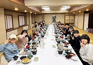 正面の床の間に掛け軸が飾られたお座敷で長く並べられたテーブルの両側に座椅子に座った20名ほどの食事の様子。