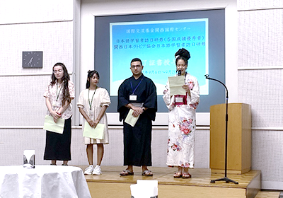 舞台上で和服を着た男性と女性、それに洋装の女性2人の計4人が並び、向かって右端の和服の組成がマイクを手に話している