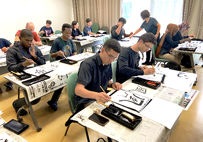 教室での習字の風景。新聞紙を敷いた机の上に半紙を広げて練習している。