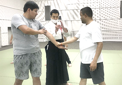 道場で合気道の道着を着た講師からレクチャーを受ける参加者の短パンTシャツ姿の2名の男性
