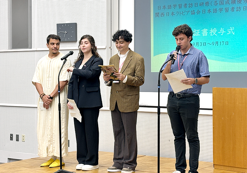 会場の前方に設置された仮説ステージに四人の人物が立ち、2本のスタンドマイクに向かって発言している