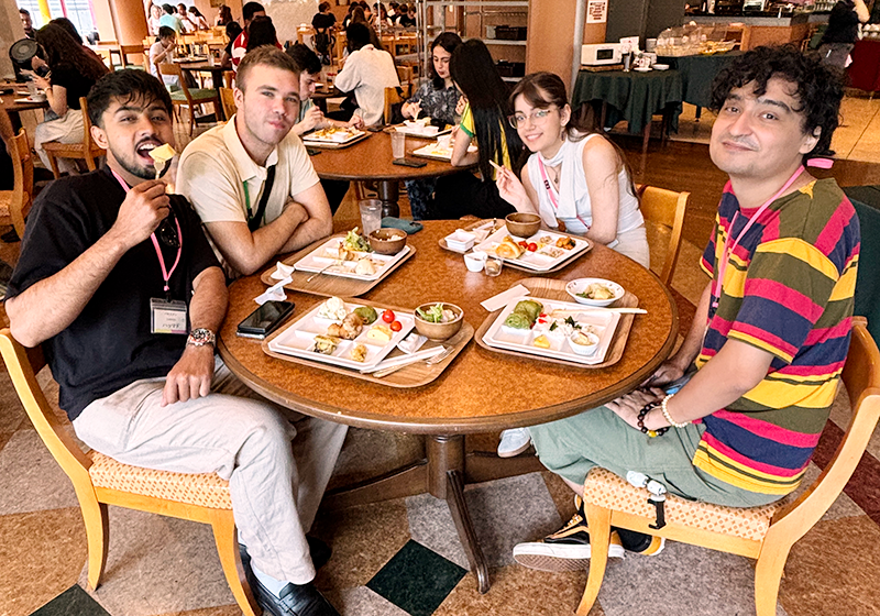 食堂で、画面手前の丸テーブルで、四人の人物がいっしょに食事をしている。