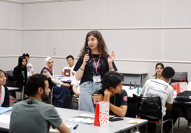 会場で一人の女性が机の間に立ちマイクで発言している。他の参加者は座って話を聞いている。