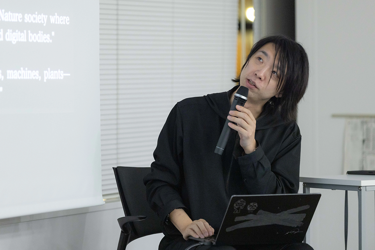 マイクを手に、画面を見上げながら話している