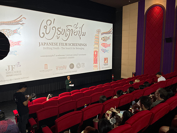 映画館にて、1人がスクリーンの前に登壇してマイクを持って喋っている。座席には15人以上来場者が座っている。