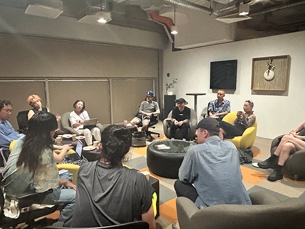A group of about 10 people are sitting around a central table in a room, conversing in chairs lined up along the wall.