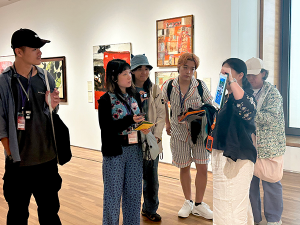 Six people gathered and having a conversation in an exhibition space with white walls decorated with paintings.