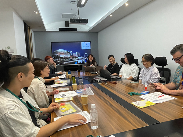 白い壁の会議室で10人ほどの参加者が向かい合って座りディスカッションしている