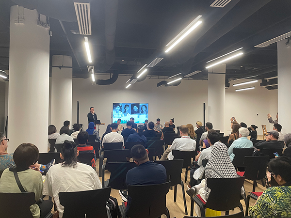 白い壁の広い会場に並べられた椅子に参加者が座って前方のモニターを見ている