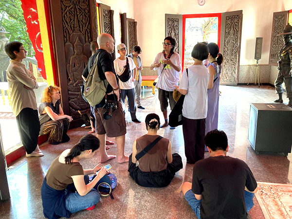 A room with open windows and doors where several people are standing or sitting in a circle chatting.