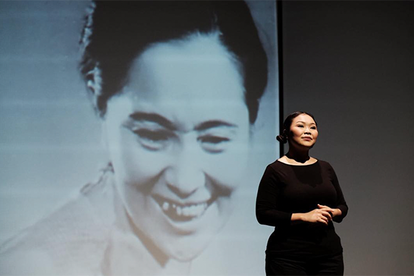 背景にモノクロ映像が写された舞台で演者が立っている