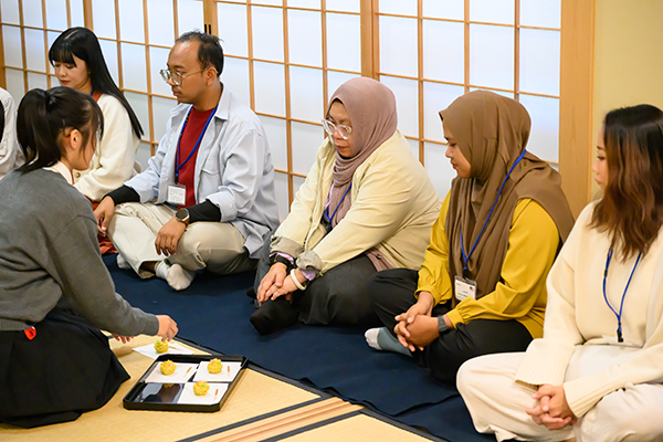 広島工業大学付属高校の茶道室で茶道を体験する参加者5名と、和菓子を提供している1人の学生。