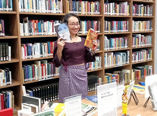 A smiling woman holding books in both hands in front of a wall-length bookshelf