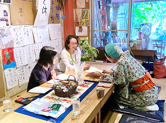 In a Japanese-style room, two women in Western clothes can be seen on either side of a wooden table, and the back of a woman in kimono can be seen in the foreground.