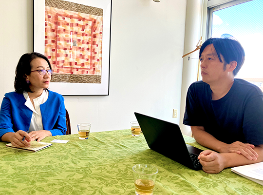 A woman wearing glasses and a blue jacket and a man wearing a black T-shirt are having a conversation at a table with a green tablecloth