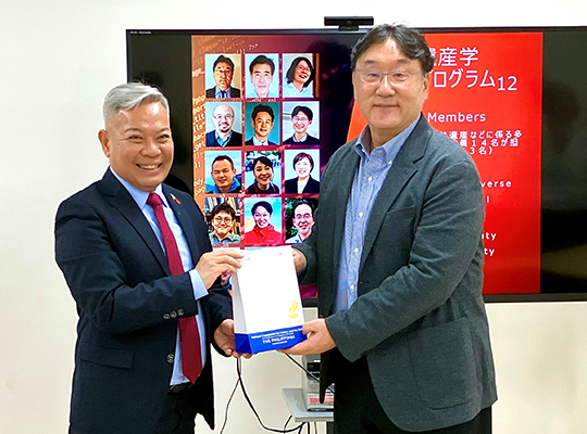 Two men in suits are standing side by side in front of a monitor