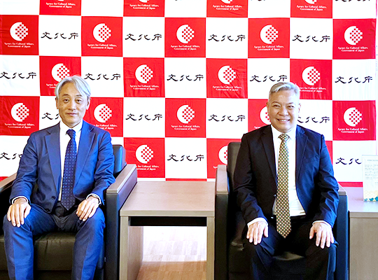 Two men sitting in chairs with the Agency for Cultural Affairs logo on the back panel in the background