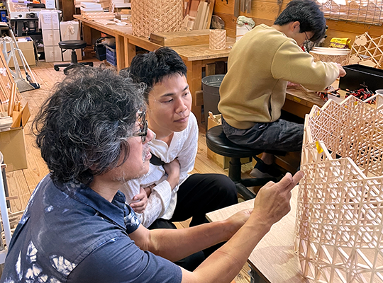 Director Pat Boonnitipat crouches down in front of a wooden craft in a corner of the workshop, listening to what the artist has to say.