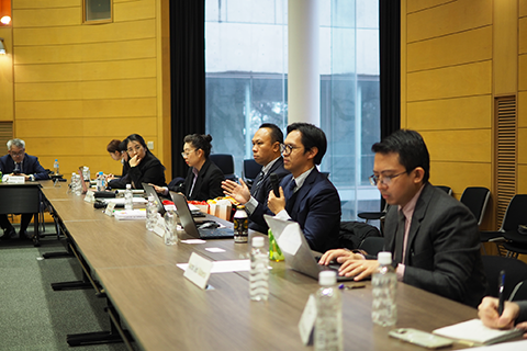 会議室のようにテーブルが組まれた場所に8人が座っている。内、1人の男性が座ったままマイクを持ち話している。