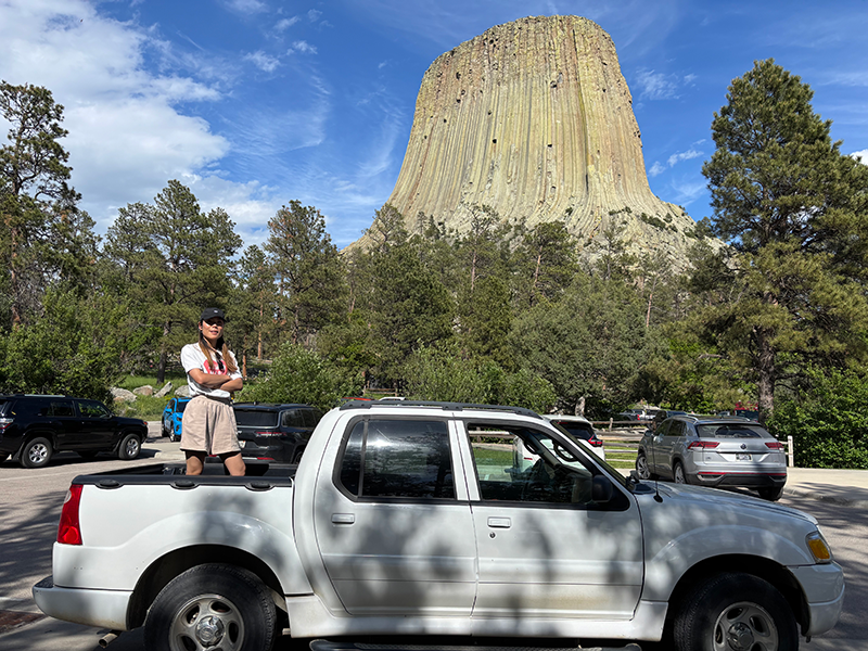 デビルズ・タワーを背景に、ピックアップトラックの荷台に乗っている高久さんが腕を組んで立っている。