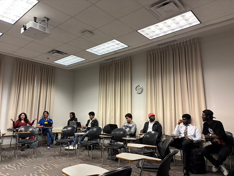 教室の壁に沿って椅子に座っている8名がディスカッションをしている。