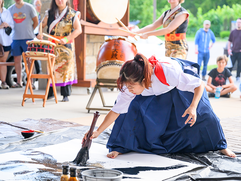 太い筆を使って力強い書道を行っている、後ろでは地元の太鼓チームが太鼓を叩いている