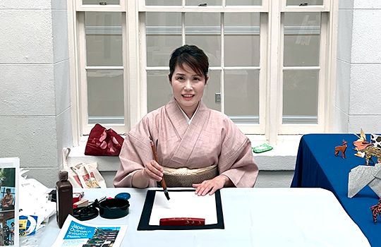 水色のカバーがかかったテーブルの前で、和服を着て半紙の前で筆を構えて準備する著者