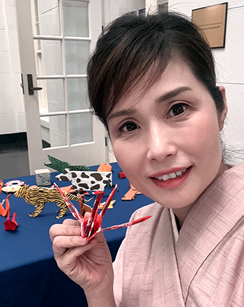 鶴や他の様々な動物たちの折り紙が並べられた机の前で、赤と白の混じった色の素材で折った鶴を片手につまんでこちらに見せている著者