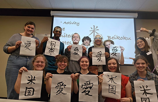 壁にかかったスライドスクリーンを背景に2列に並んだ10人ほどの人物が、愛、米国などと書かれた習字の半紙を掲げている