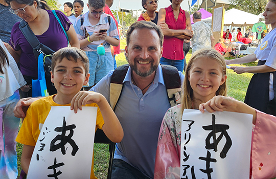 リュックを背負った男性の左に男の子、右に女の子が習字を手に掲げて笑っている