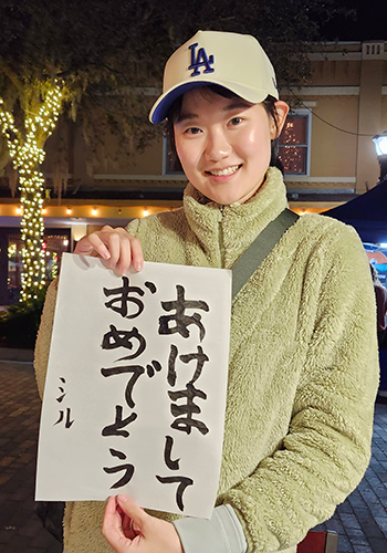 フリースを着てLAのキャップを被った女性があけましておめでとうと習字で書かれた半紙を掲げて嬉しそうにこちらを見ている
