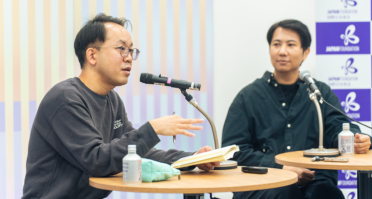 インタビューの様子：１冊の本を手に取りマイクに向かって話す福冨氏とそれを見ているチラット氏