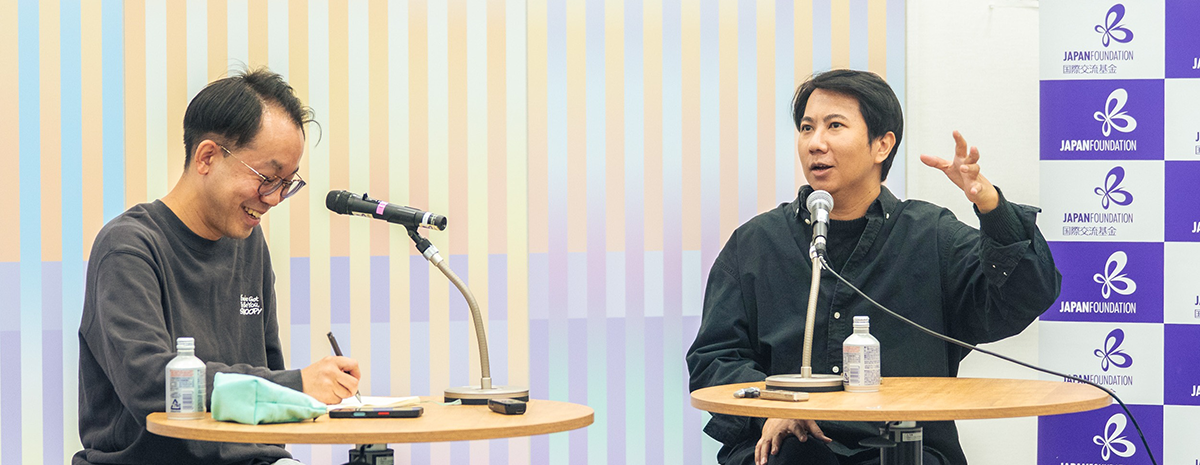 インタビューの様子：何かをメモする福冨氏と福冨氏に話しかけるチラット氏