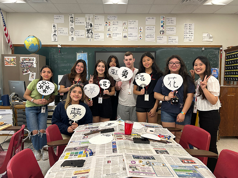 教室にて、男女9名が漢字が書かれているうちわを持って写っている。