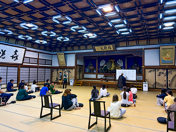 福井県永平寺町にある大本山永平寺の坐禅体験を行っている大広間に参加者20名と1名司会者のような人物とお坊さんがいる。