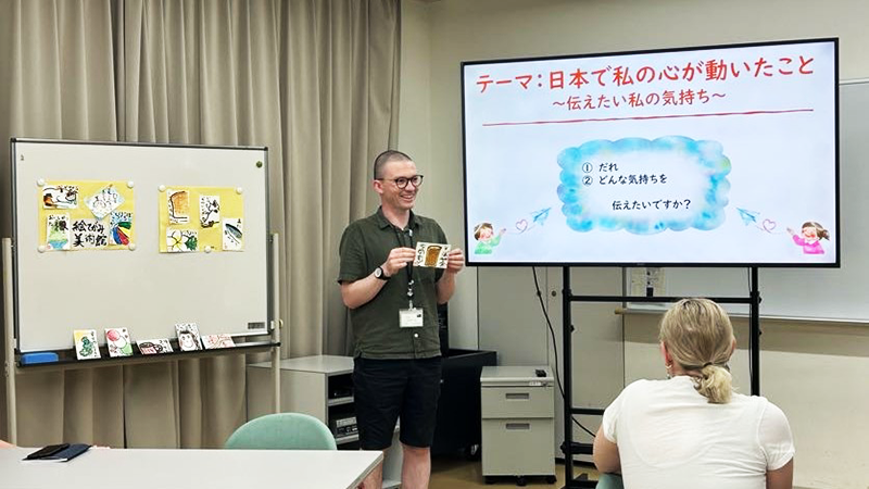 教室の右側には参加者の作品が貼られたホワイトボードがあり、左側には絵手紙のテーマ「日本で私の心が動いたこと」がモニターに映し出されていて、一人のニュージーランドの日本語教師の参加者が、他の参加者の前で自分の作品について語っている。