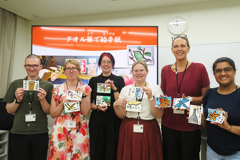 ニュージーランドの日本語教師6人の参加者がそれぞれ自分の絵手紙作品を手にもって見せている写真。絵手紙にはもみじ、とんぼ、風鈴、街並み、湯飲み等の絵がかかれ、「一緒に日本に行こう」「涼しい風」「歩こう」などの短い文が添えられている。